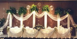 DECORATION by Wedding Specialties, Orlando — This Wedding Backdrop will frame your wedding party photos using a combination of traditional Roman columns with lights, greens, balloon bubbles and elegant fabric drapery on columns and table front.